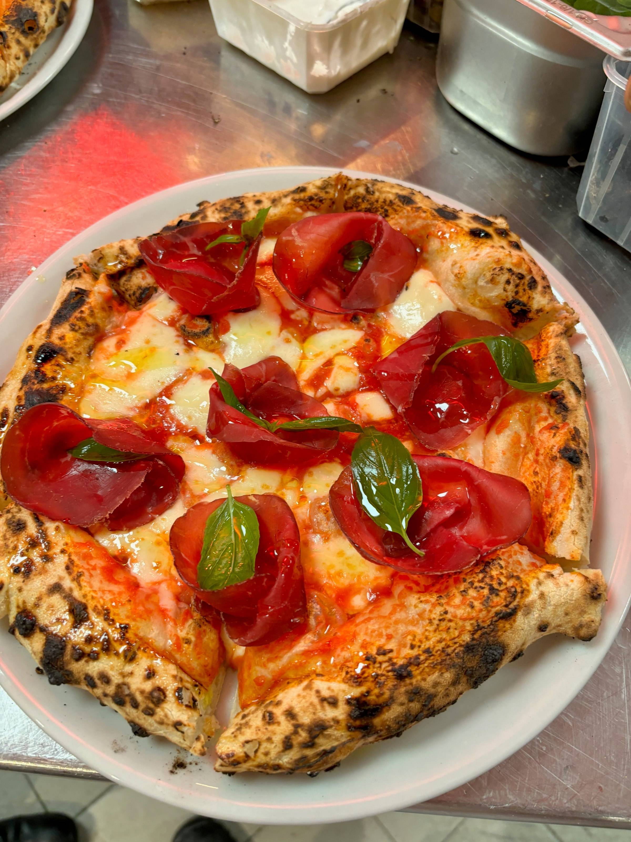 Pizza à la bresaola posée sur le plan inox du poste, sous la lampe chauffante, entre les bacs de mise en place.
