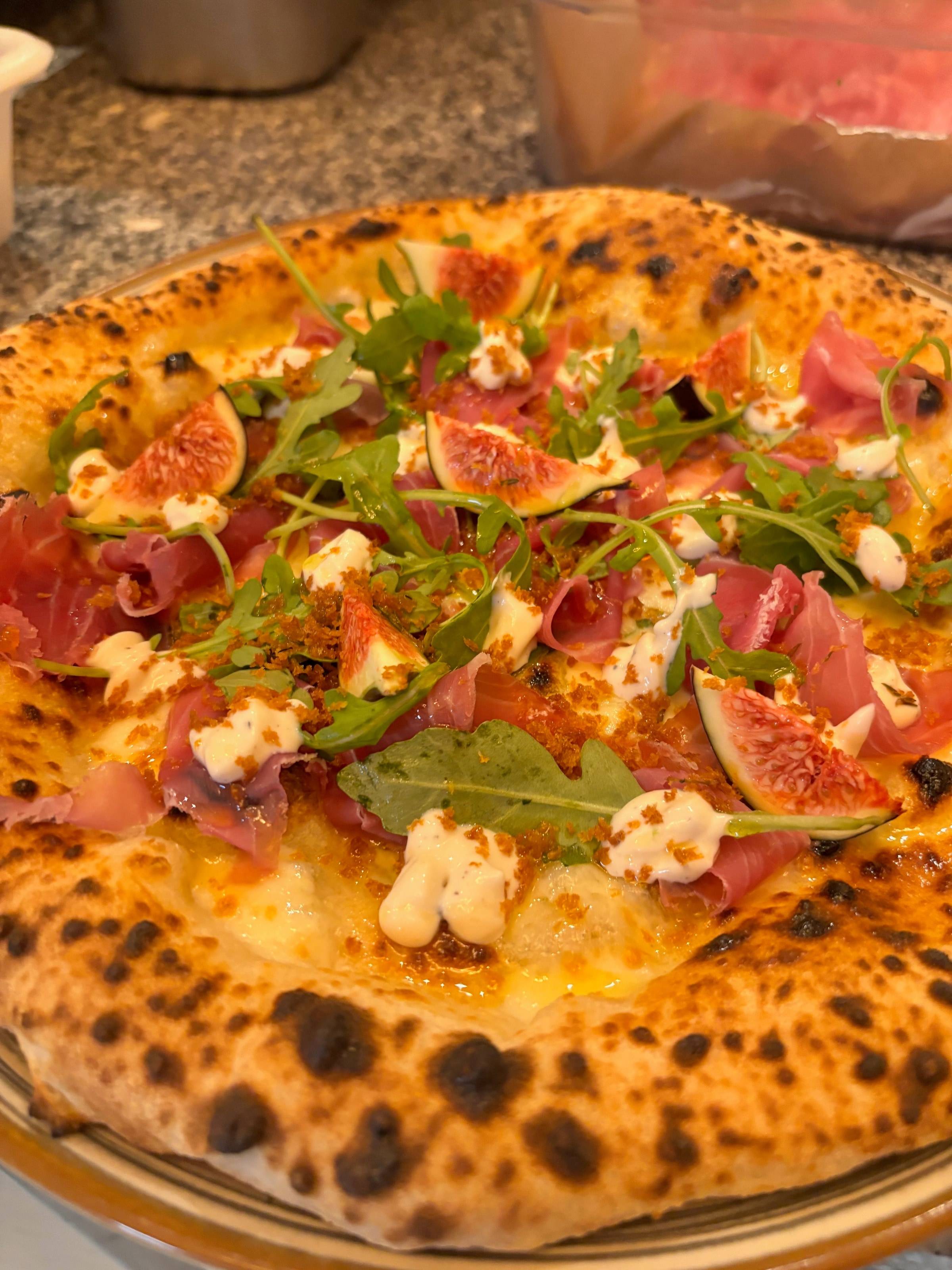 Gros plan de trois-quarts sur une pizza garnie de figues, de jambon cru et de roquette, cornicione léopardé au premier plan.