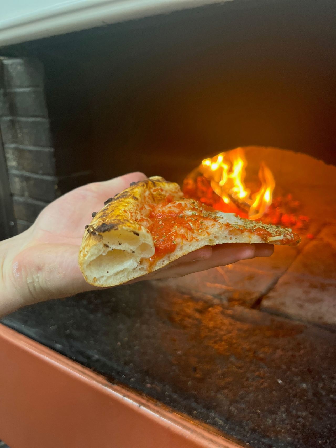 Une part de pizza tenue à plat dans la main devant les flammes d'un four à bois : on voit l'alvéolage du cornicione.