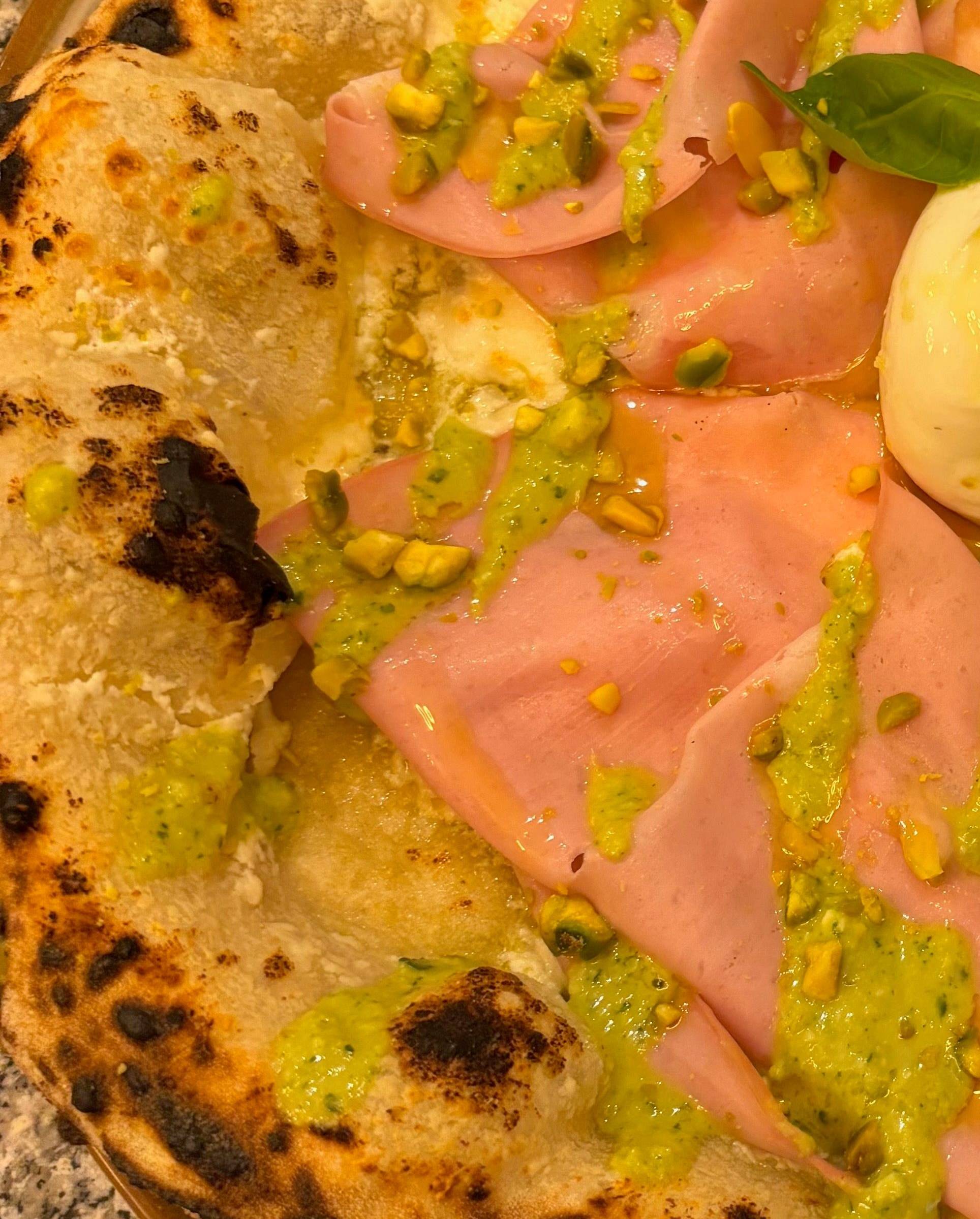 Gros plan sur un cornicione gonflé et léopardé, au bord d'une pizza à la mortadelle et au pesto de pistache.