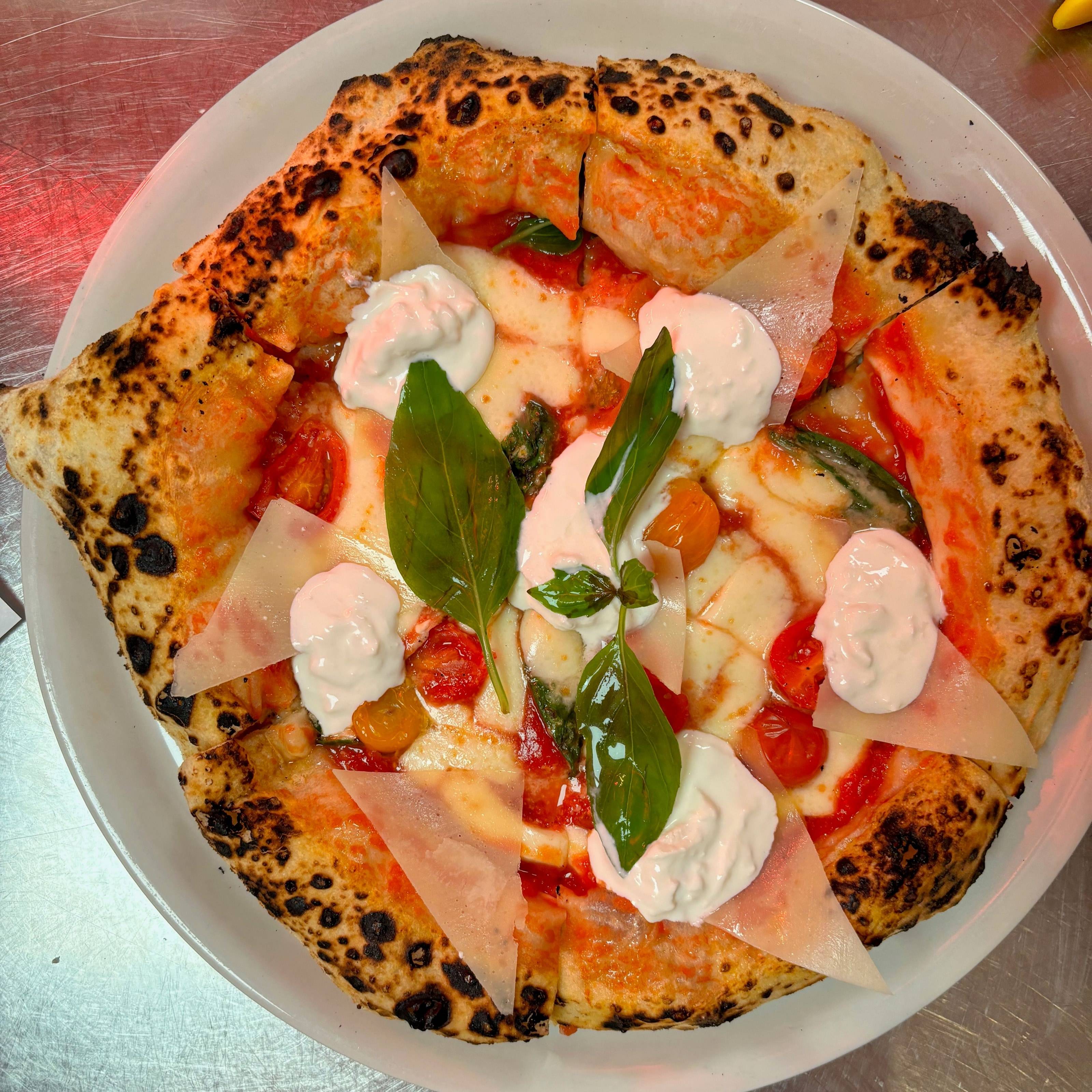 Pizza vue de dessus avec des tomates cerises, des cuillerées de stracciatella, des copeaux et de grandes feuilles de basilic.